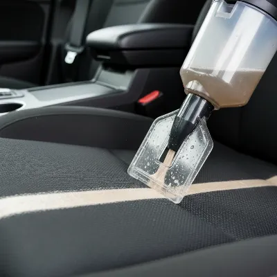 A portable carpet cleaner being used to clean a dirty fabric car seat, with an extension hose reaching into the car. A person is holding the cleaning tool, demonstrating the ease of use and the machine's compact design.